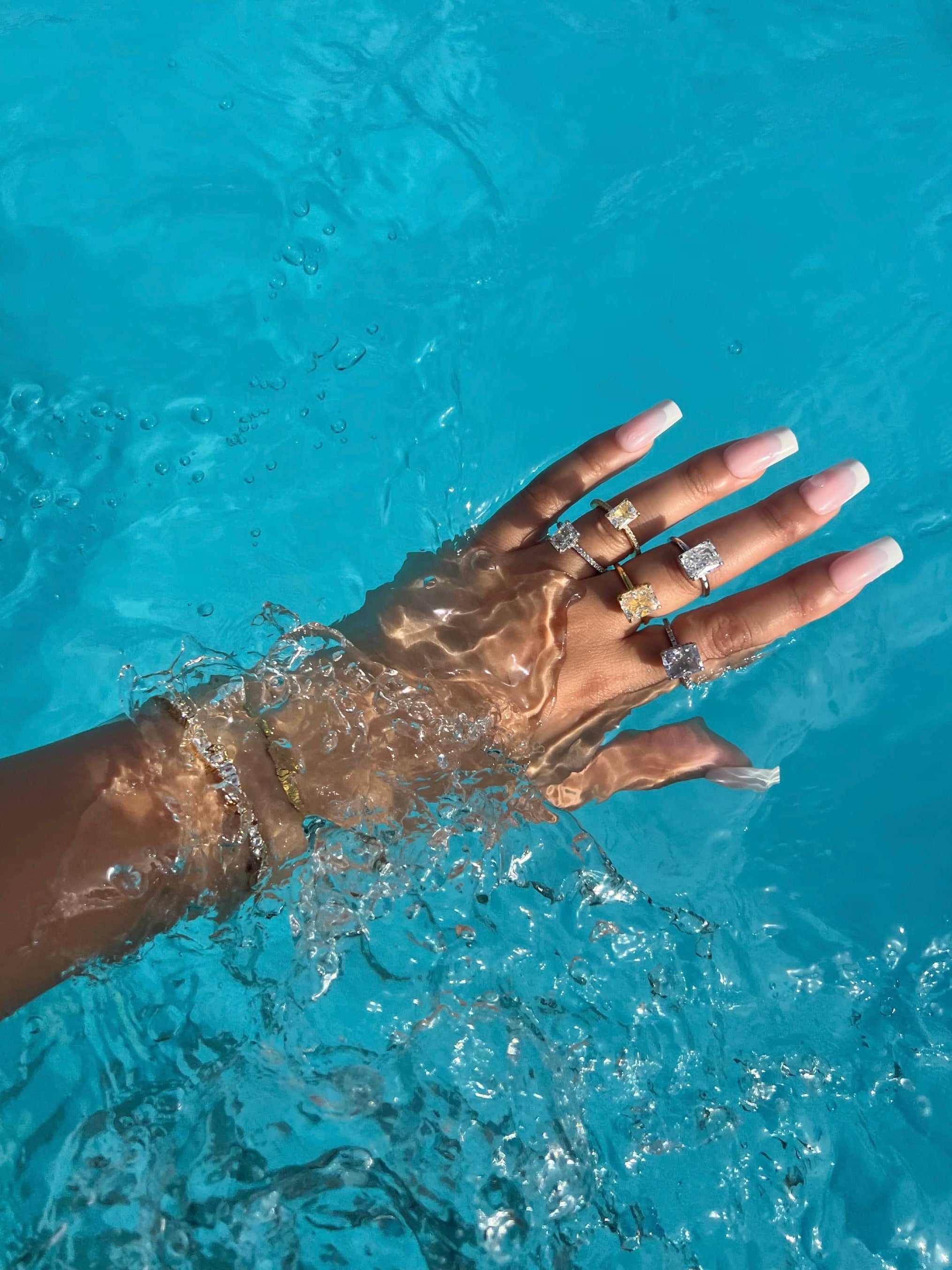 A woman’s hand adorned with five rings, three featuring large silver cubic zirconia stones on a silver band, and two on a gold band, rests gracefully in a blue pool of water filled with bubbles. The hand also showcases elegant white French manicured nails