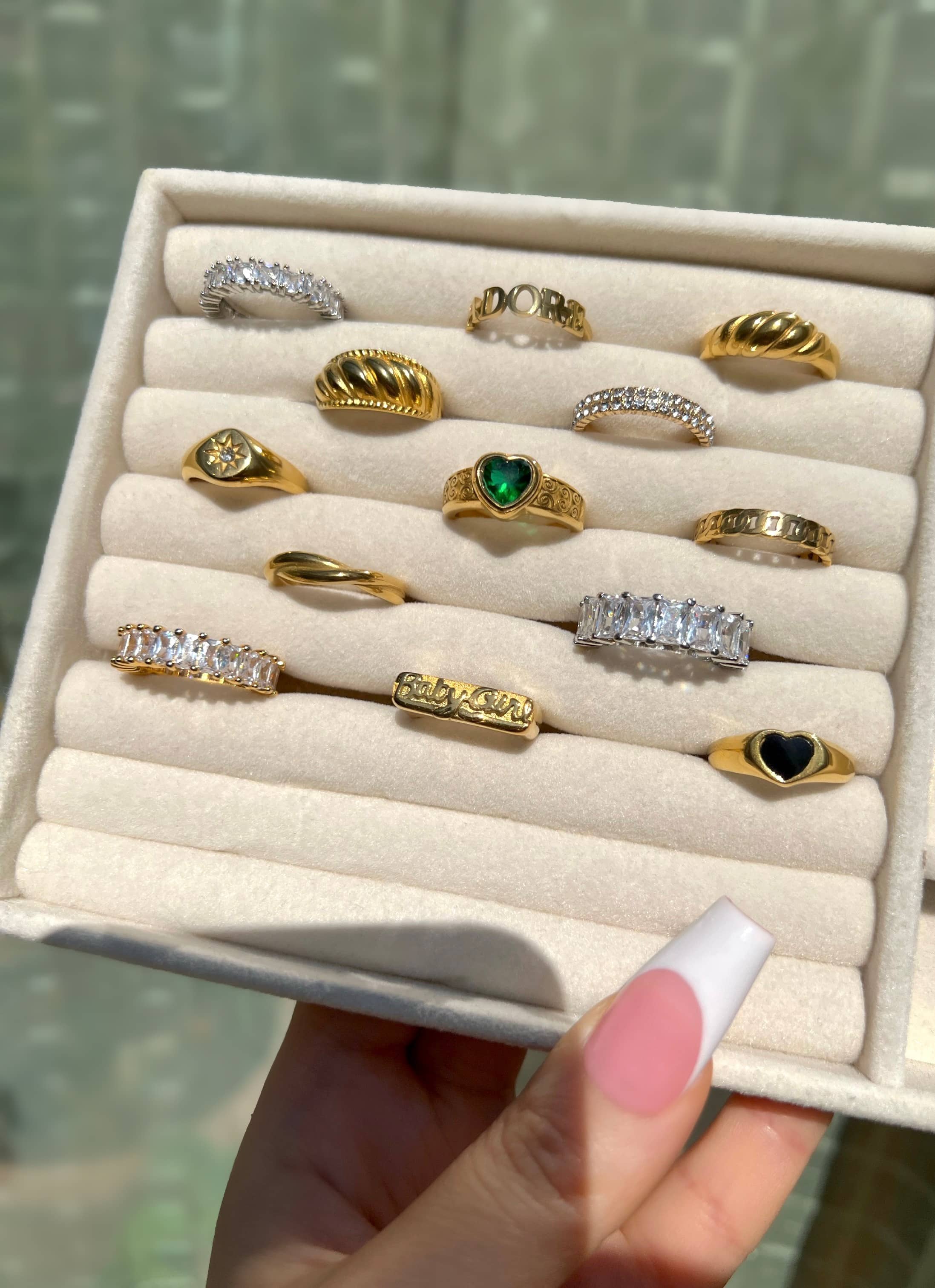 A woman’s hand with french manicured nails holds a white ring holder displaying 13 rings. The rings feature diamond simulant rings, a green heart-shaped stone, black heart ring, croissant rings, baguette eternity rings, baby girl ring