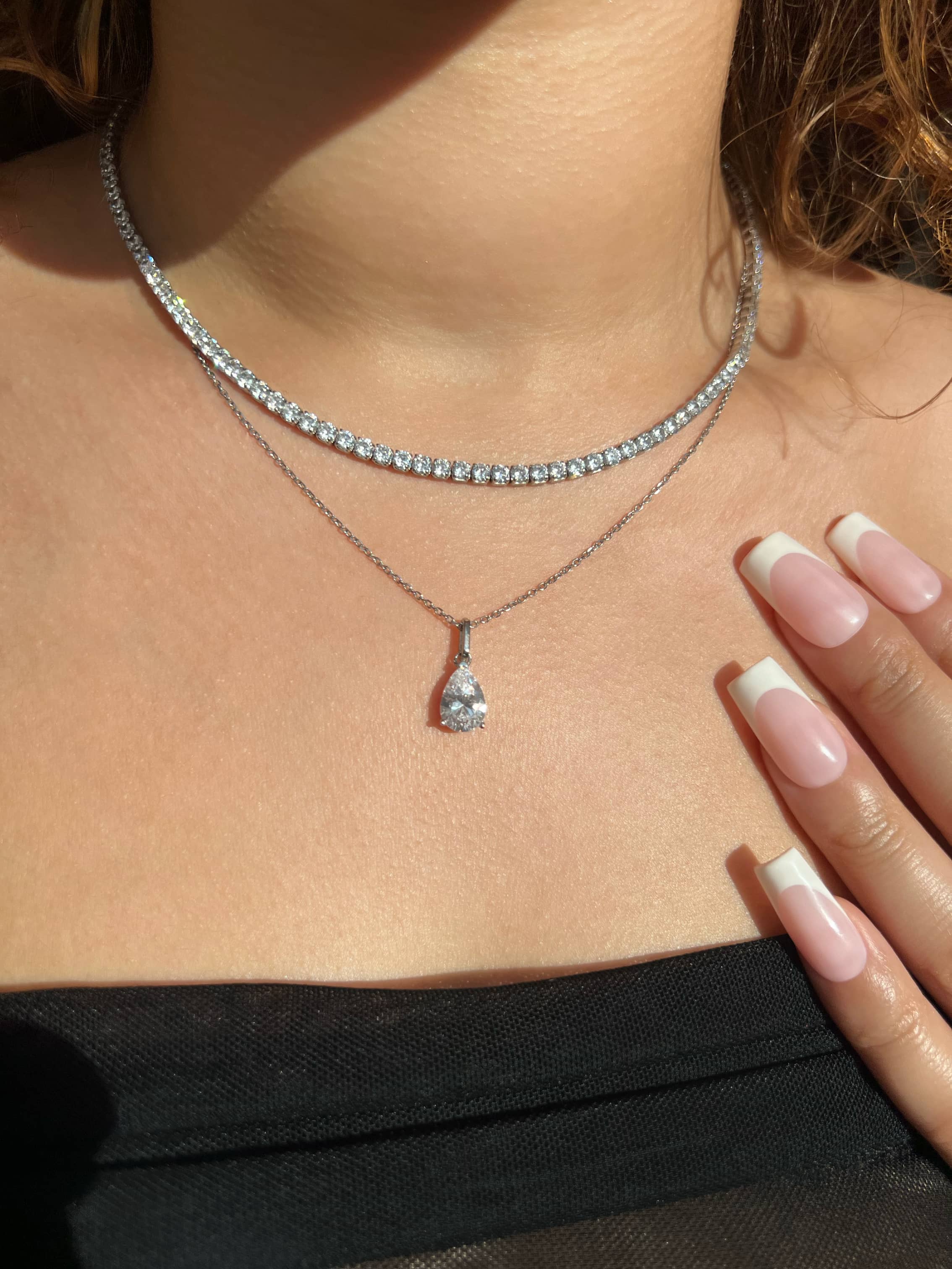 A close-up of a woman’s chest adorned with a silver tennis diamond necklace paired with a stunning silver teardrop-shaped pear cut pendant necklace, with her hand elegantly on her chest with French manicured nails. She is wearing a black bandeau mesh top