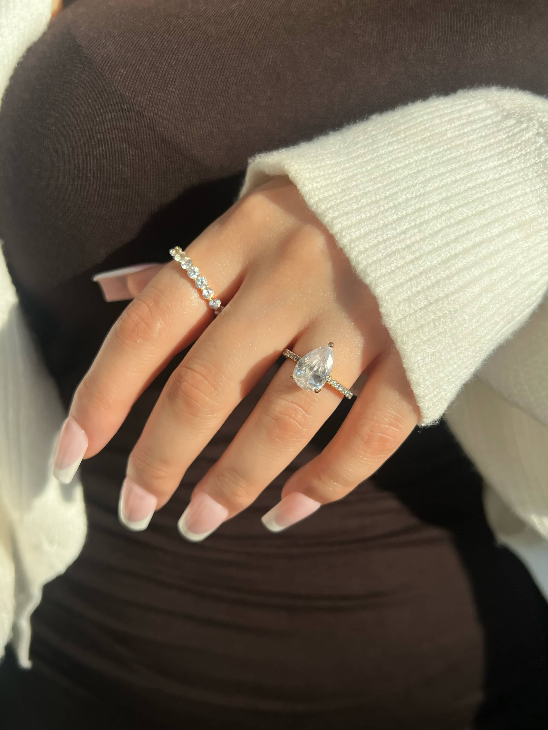 A woman's hand with elegant French manicured nails showcases a beautiful gold band featuring a stunning pear-shaped diamond. The hand also wears a second round cut row of smaller diamonds in a band. She is wearing a brown dress with a white cardigan