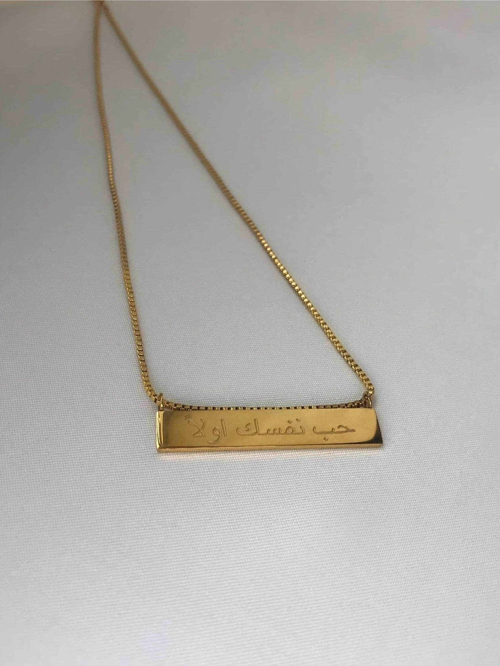 A gold chain necklace featuring a rectangular pendant inscribed with Arabic writing that says "حب نفسك اولا," translating to "Love yourself first." The elegant design highlights the beauty of the necklace, making it a meaningful accessory for any occasion