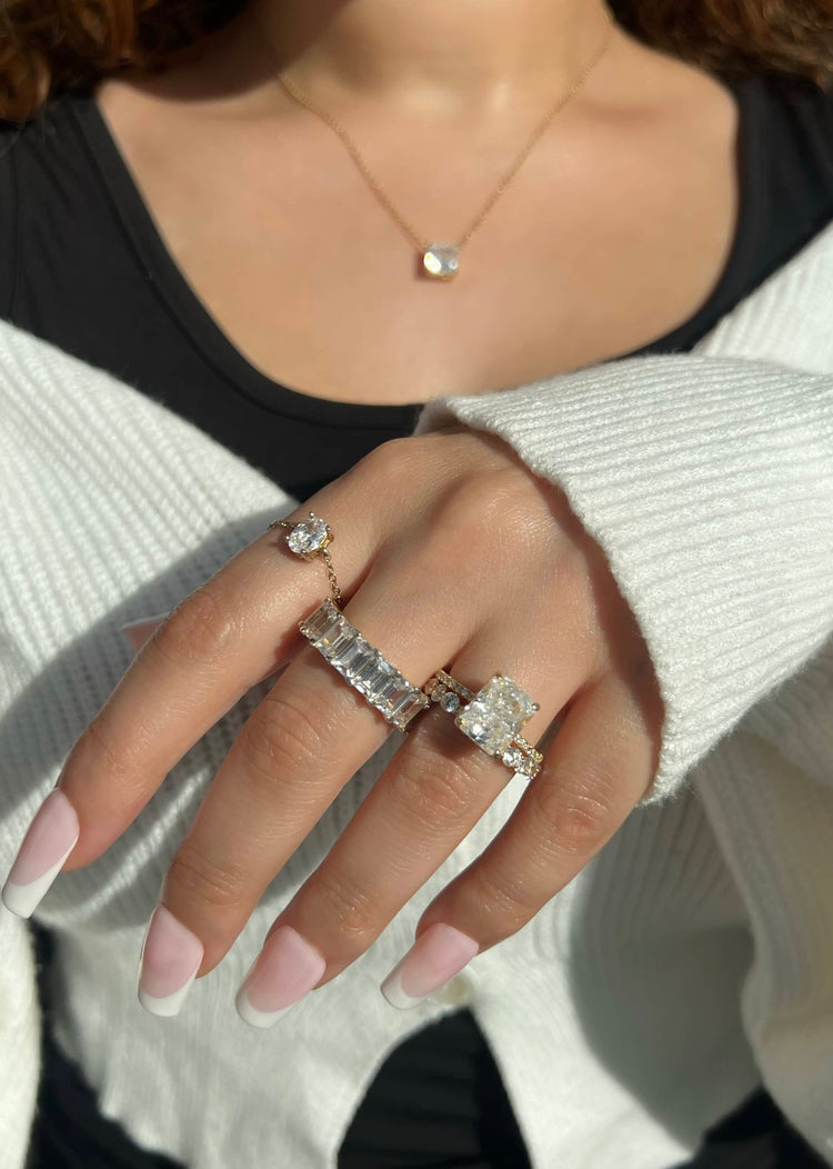 A woman’s hand adorned with four stunning gold diamond rings: a thick emerald cut band, a round cut band, a large radiant-cut diamond, and a small chain ring. The hand also features a gold chain with a diamond pendant,
complemented by a French manicure. 