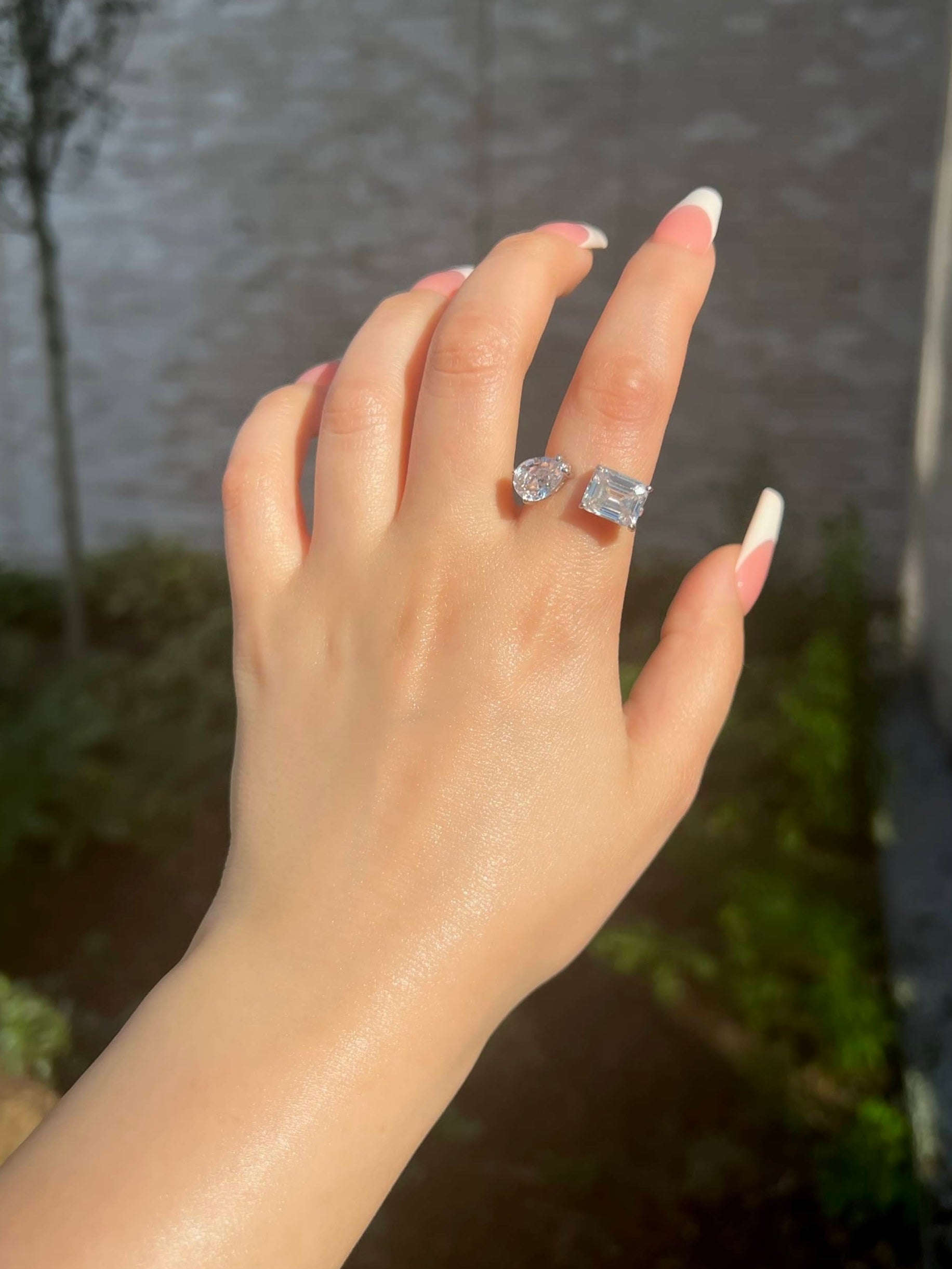 A woman’s hand showcasing a striking silver ring featuring two large, sparkling gemstones: a pear-shaped stone on the left and a square-shaped stone on the right. The hand has elegantly French-manicured nails, adding sophistication to the overall look 