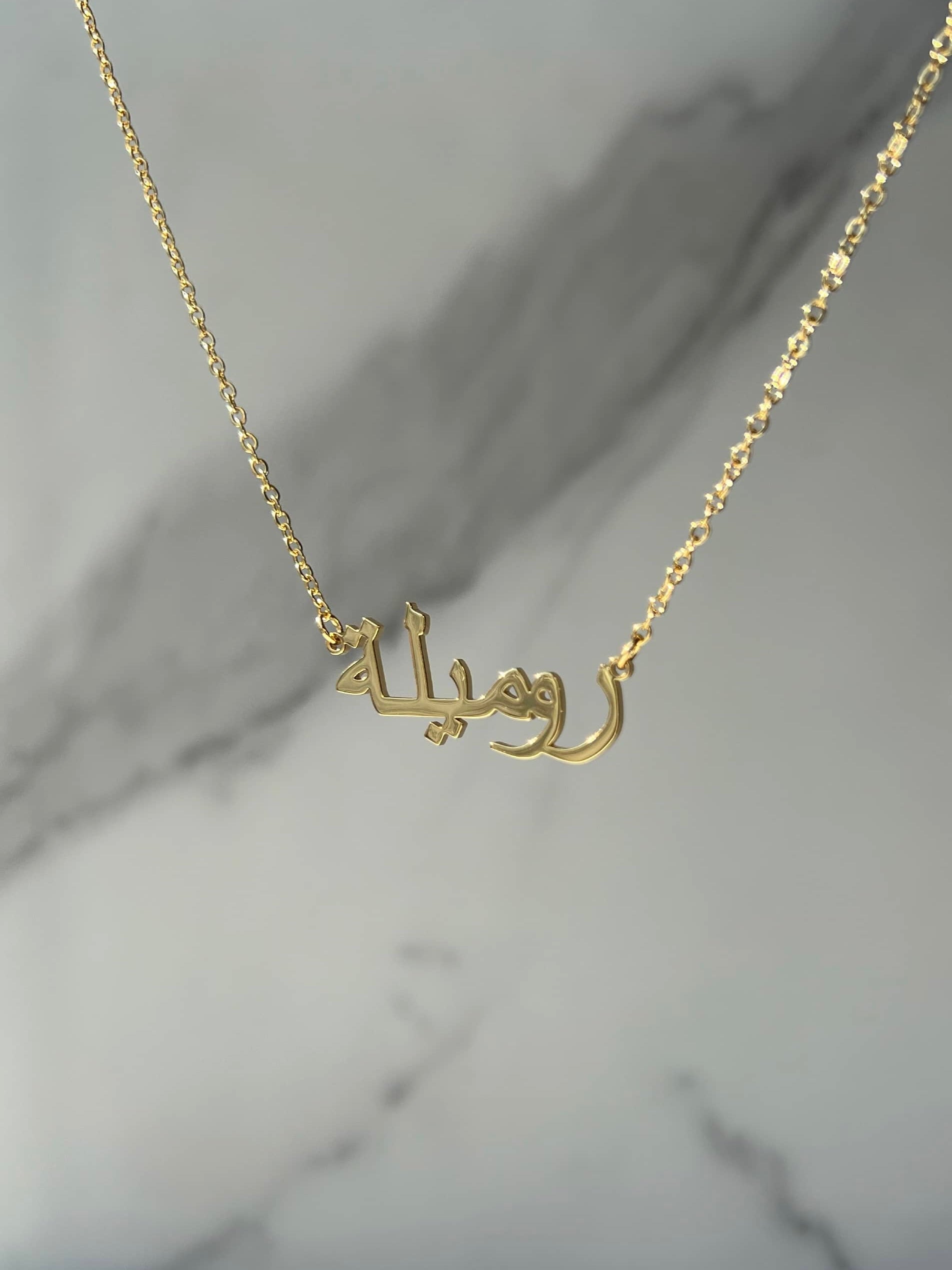 A beautiful gold necklace featuring a pendant with intricate Arabic calligraphy translating to ‘Romeela’. The delicate gold chain complements the pendant, creating an elegant look. The necklace is showcased on a grey marble drop background 
