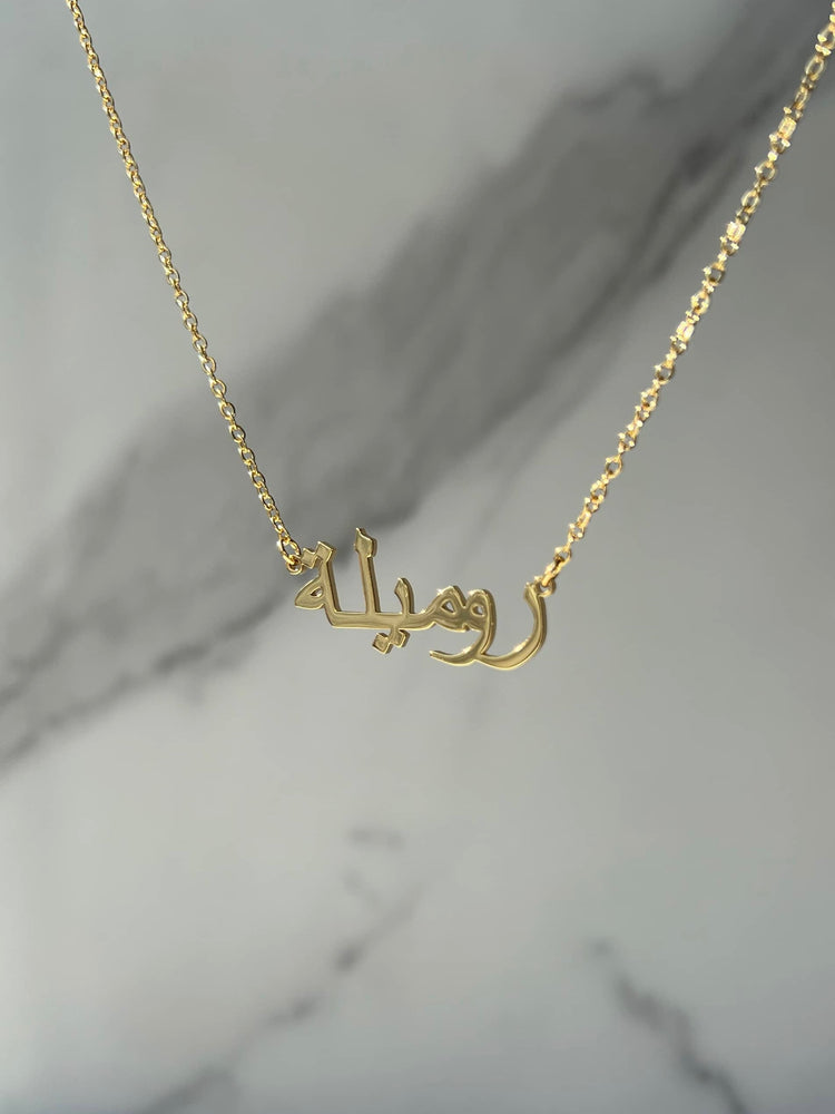 A beautiful gold necklace featuring a pendant with intricate Arabic calligraphy translating to ‘Romeela’. The delicate gold chain complements the pendant, creating an elegant look. The necklace is showcased on a grey marble drop background 
