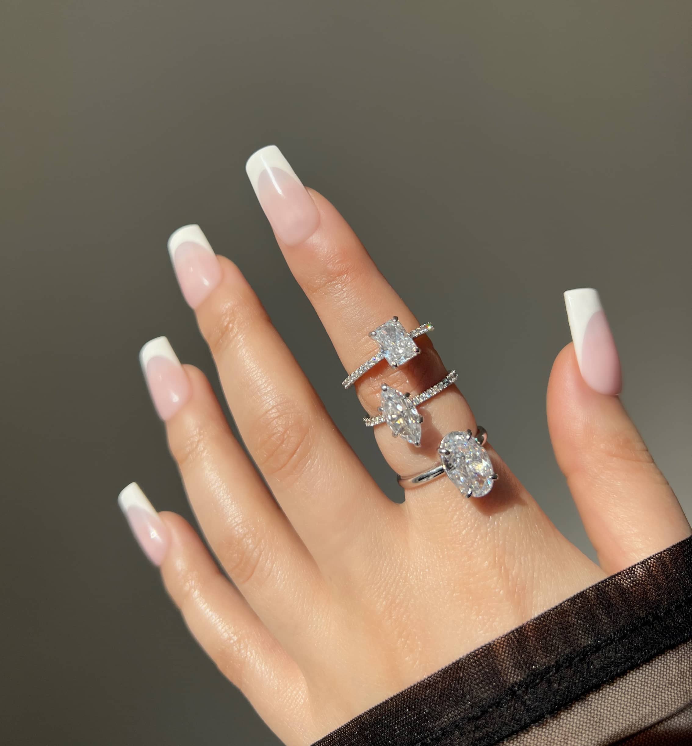 A woman’s hand with French manicured nails displays three silver rings featuring different shaped clear stones: a rectangular stone, a marquise stone, and an oval stone, showcasing a stylish and elegant jewelry collection
