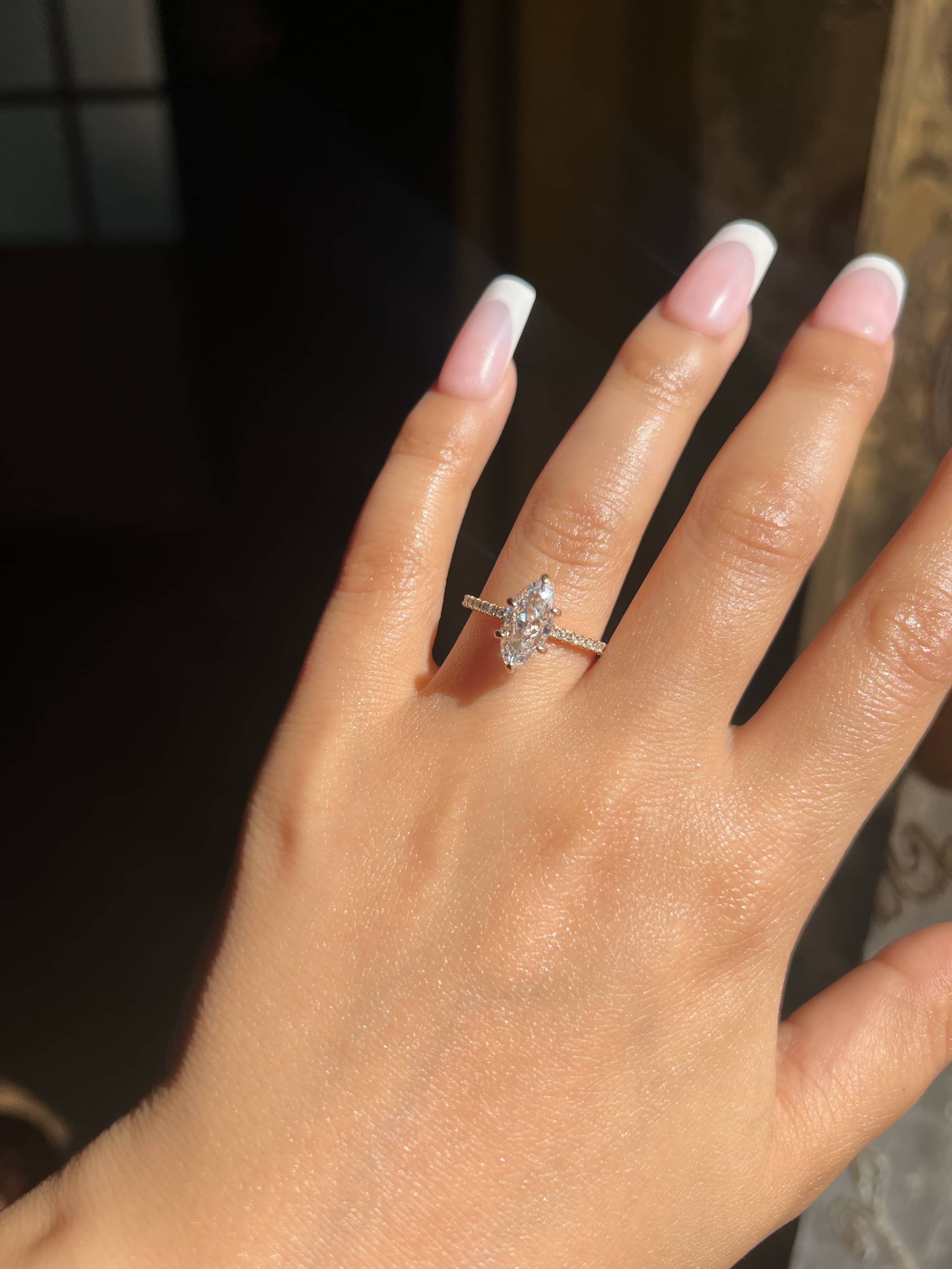 A woman’s hand displaying a stunning marquise shaped gold diamond engagement ring on the ring finger. The ring features a thin gold band adorned with small diamonds, complemented by elegantly manicured French nails