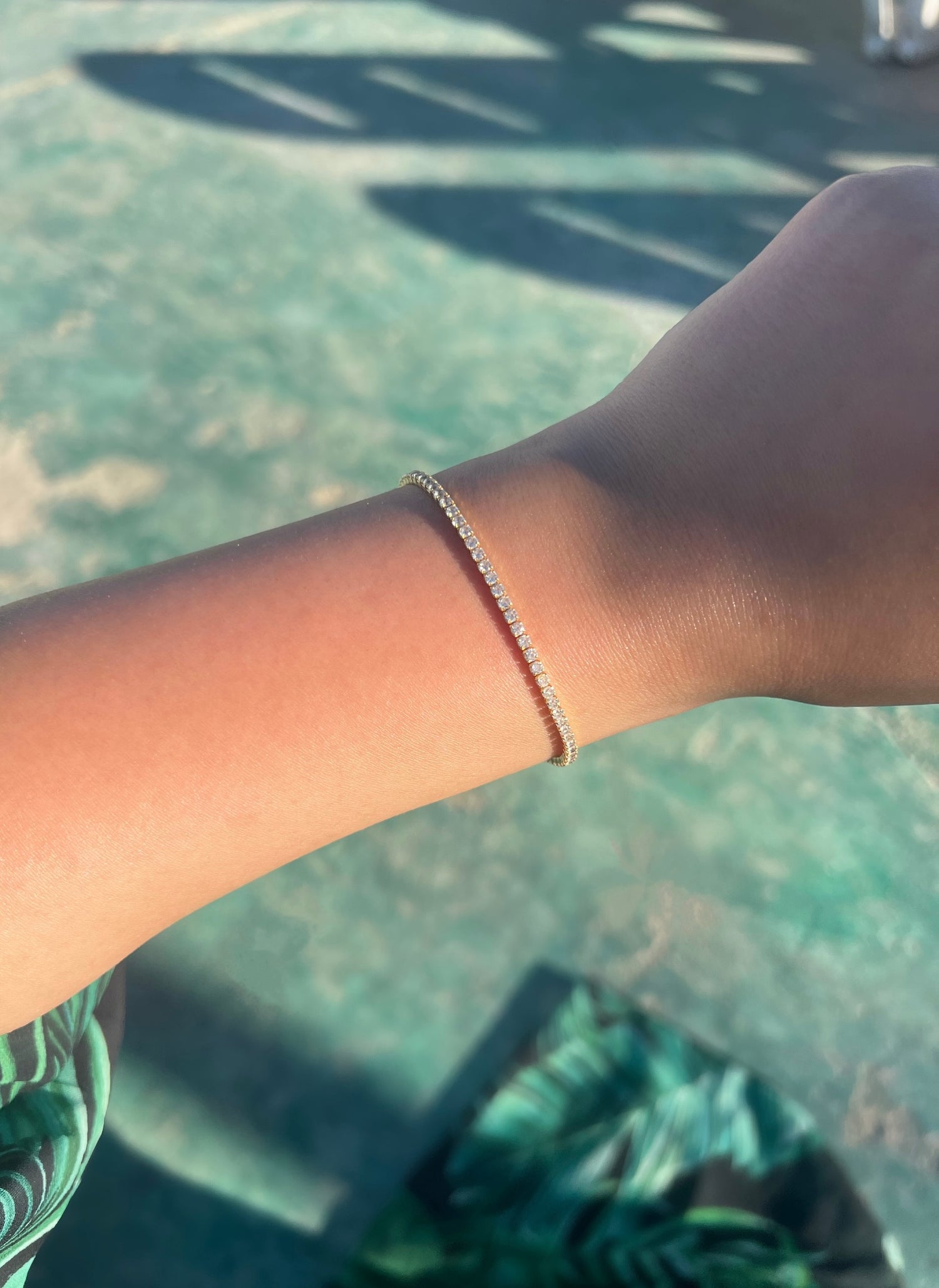A close-up of a woman’s arm adorned with a thin 2mm gold bracelet featuring small diamonds. The arm rests on a green surface with a floral pattern in shades of green and black, highlighting the elegance of the bracelet against the vibrant background. 
