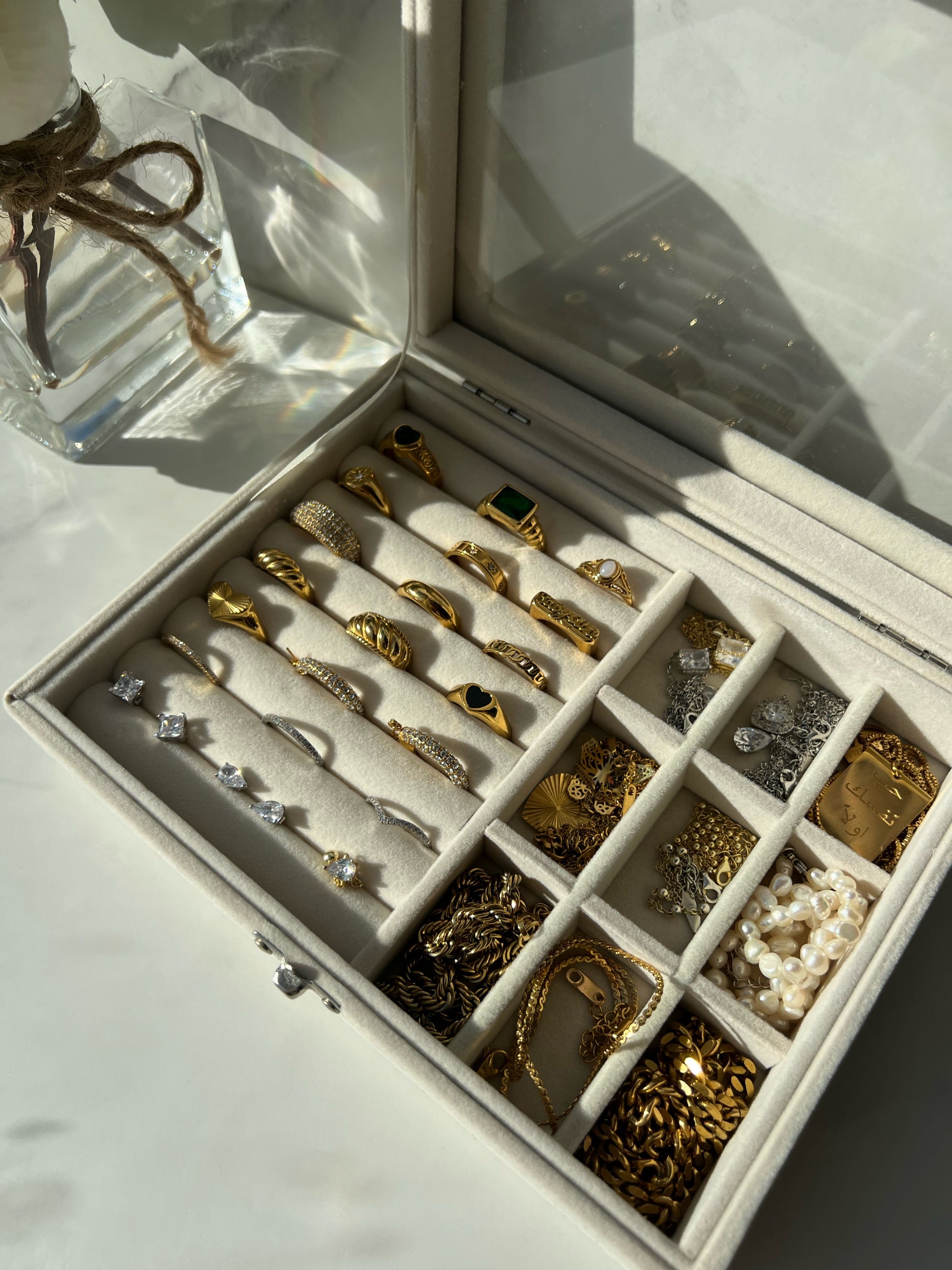 A white jewelry box with a glass lid, displaying an assortment of gold rings, earrings, gold silver, and pearl necklaces, all neatly arranged. There is a gold chain necklace featuring a large rectangular pendant with Arabic text. The focus is the rings